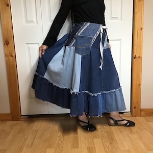 May include: A long, patchwork denim skirt featuring various shades of blue fabric, including light and dark wash panels. The skirt has a frayed hem, a visible back pocket from a pair of jeans, and a white ribbon tie closure at the waist.
