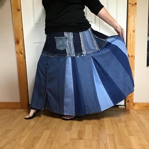 May include: A floor-length denim skirt featuring a patchwork design with various shades of blue. The upper section includes a small pocket, while the lower portion consists of alternating vertical panels of light and dark blue denim fabric.