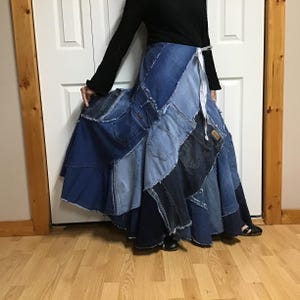May include: A floor-length skirt constructed from various shades of blue denim fabric patches. The garment features multiple visible back pockets from jeans, raw frayed edges, and a white ribbon tie closure at the waist, creating a full silhouette.