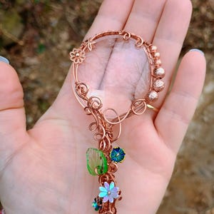 May include: A copper-colored wire-wrapped magnifying glass pendant with a clear glass lens. The pendant features decorative floral and leaf accents in green, blue, and iridescent colors. The handle is adorned with small copper beads and a flower.