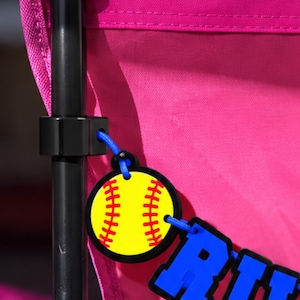 May include: A bright pink folding chair featuring a black metal frame and a hanging decorative tag. The tag consists of a yellow softball with red stitching and a blue name plate with the letters RIL, attached by a blue cord to the chair frame.