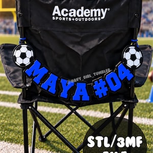 May include: A black folding chair with the Academy Sports + Outdoors logo on the backrest. Attached to the chair is a blue and black custom nameplate reading MAYA #04, flanked by two circular black and white soccer ball icons on each side.