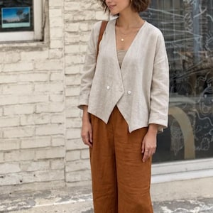 Puede incluir: Un blazer beige de doble botonadura con mangas tres cuartos y dos botones blancos, usado sobre una parte superior beige a juego. El conjunto se combina con pantalones anchos de cintura alta en color óxido y zapatos planos negros.