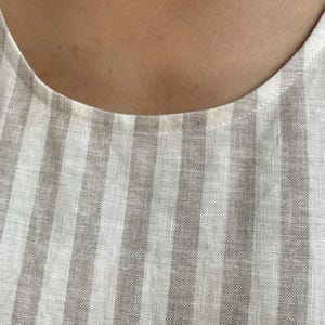 May include: A sleeveless top featuring a vertical striped pattern in alternating shades of light beige and white. The garment has a simple round neckline with a clean, finished hem along the collar, showcasing a woven fabric texture throughout.