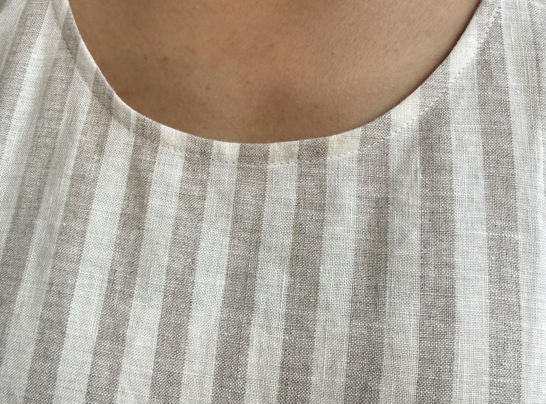 May include: A sleeveless top featuring a vertical striped pattern in alternating shades of light beige and white. The garment has a simple round neckline with a clean, finished hem along the collar, showcasing a woven fabric texture throughout.