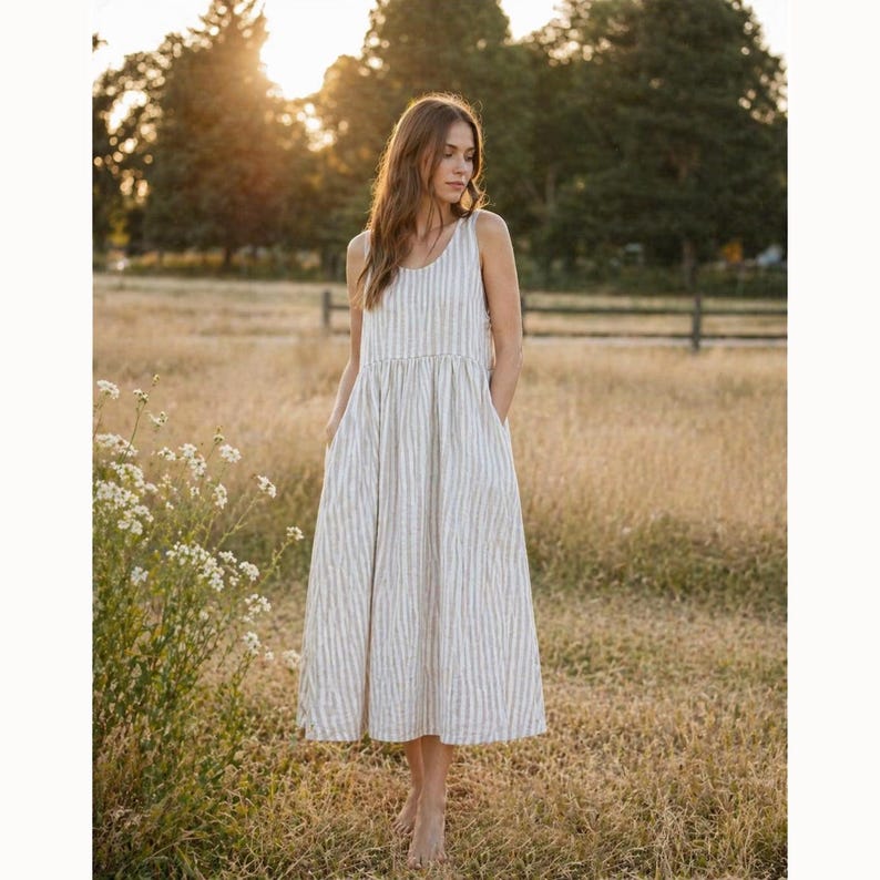 May include: A sleeveless midi dress featuring a vertical striped pattern in neutral beige and white tones. The garment has a round neckline, a fitted bodice that transitions into a gathered waistline, and a flowing skirt with side pockets.