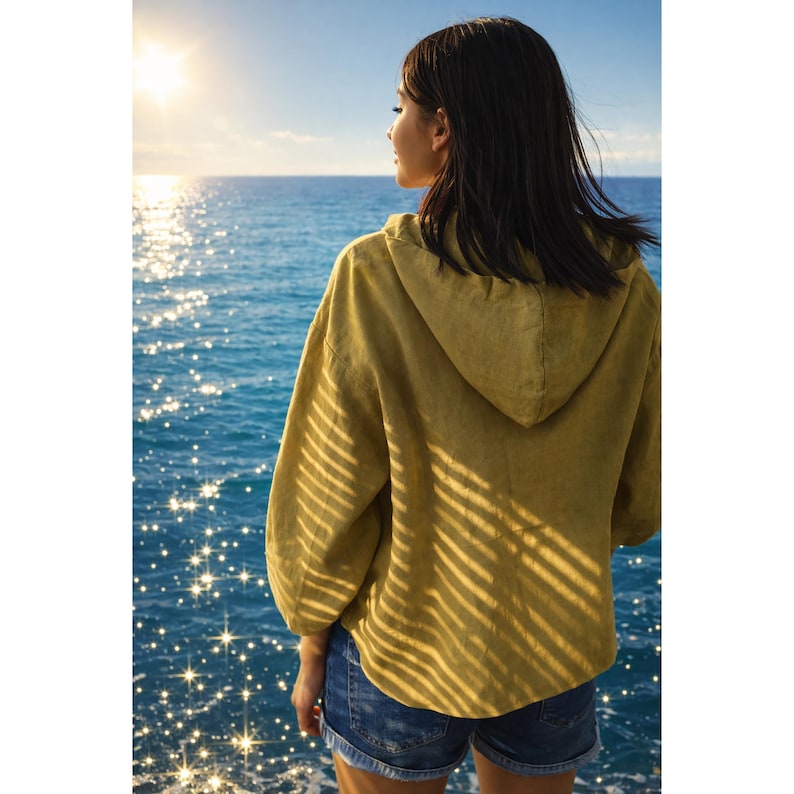 May include: A woman wearing a mustard-colored hooded jacket and denim shorts stands near the ocean, facing the horizon. The sun shines brightly, reflecting off the water. The jacket has long sleeves and a hood.