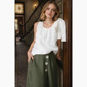 May include: A woman wearing a white linen top with a scoop neckline and a green skirt with large white buttons. The top has a ruffled sleeve detail. The skirt is a solid olive green color. The outfit is casual and stylish.