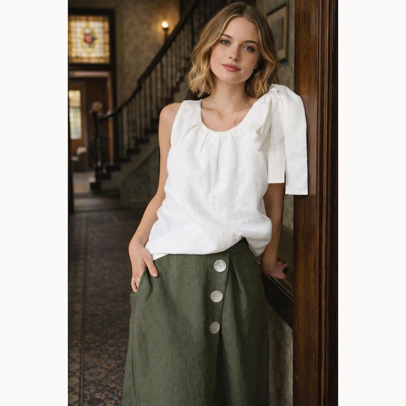May include: A woman wearing a white linen top with a scoop neckline and a green skirt with large white buttons. The top has a ruffled sleeve detail. The skirt is a solid olive green color. The outfit is casual and stylish.