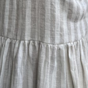 May include: A garment featuring a vertical striped pattern in alternating shades of off-white and light beige. The fabric has a visible woven texture and includes a horizontal seam across the middle, where the lower section is gathered into pleats.