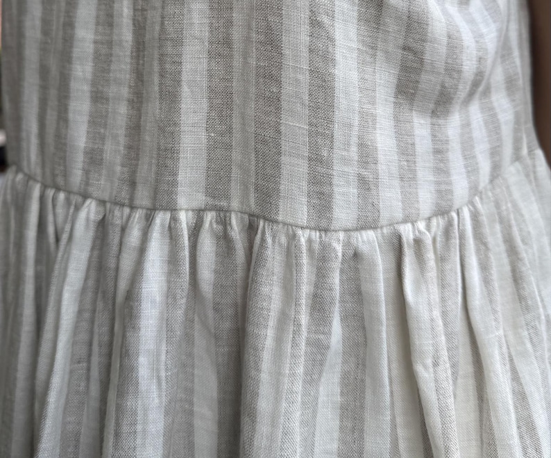 May include: A garment featuring a vertical striped pattern in alternating shades of off-white and light beige. The fabric has a visible woven texture and includes a horizontal seam across the middle, where the lower section is gathered into pleats.