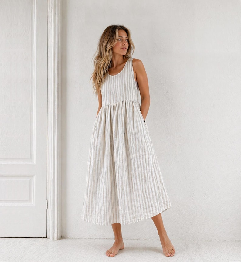 May include: A sleeveless midi dress featuring a vertical striped pattern in off-white and beige. The garment has a round neckline, a fitted bodice that transitions into a gathered, voluminous skirt, and side pockets, falling to mid-calf length.