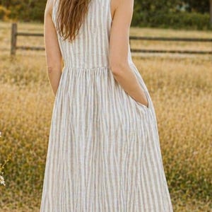 May include: A sleeveless midi dress featuring a vertical striped pattern in shades of beige and white. The garment has a fitted bodice, a gathered waistline, and a full, flowing skirt that includes side pockets, providing a simple and functional design.