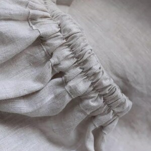 May include: Close-up of a light beige linen fabric with gathered detailing. The fabric appears soft and textured, with visible weave patterns. The gathered section creates a ruffled effect, adding visual interest to the textile.