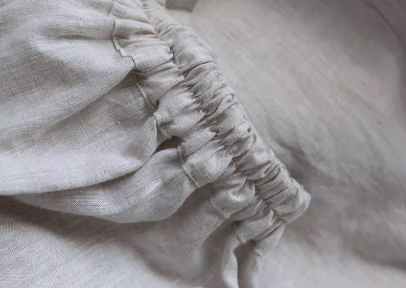 May include: Close-up of a light beige linen fabric with gathered detailing. The fabric appears soft and textured, with visible weave patterns. The gathered section creates a ruffled effect, adding visual interest to the textile.