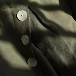 May include: Close-up of olive green fabric with three large, round, pearlescent buttons. The fabric has a textured appearance, and the buttons are evenly spaced down the front. The image is lit with natural light, creating shadows.