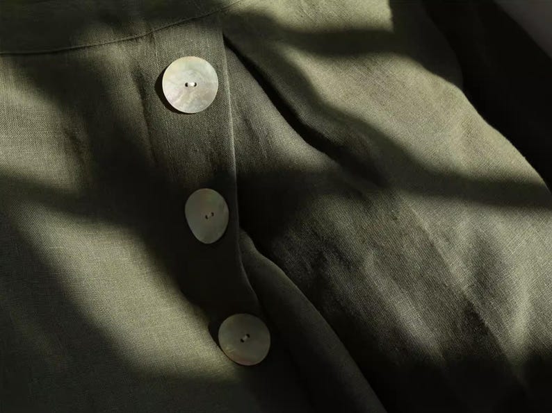 May include: Close-up of olive green fabric with three large, round, pearlescent buttons. The fabric has a textured appearance, and the buttons are evenly spaced down the front. The image is lit with natural light, creating shadows.