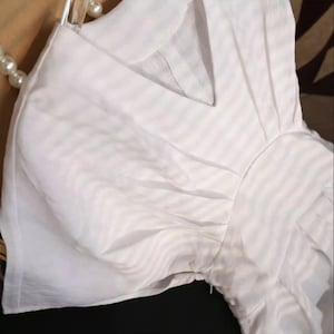 May include: A white, short-sleeved blouse with a V-neckline. The fabric has a subtle striped pattern. The blouse is hanging on a clear hanger, with a pearl necklace visible in the background.