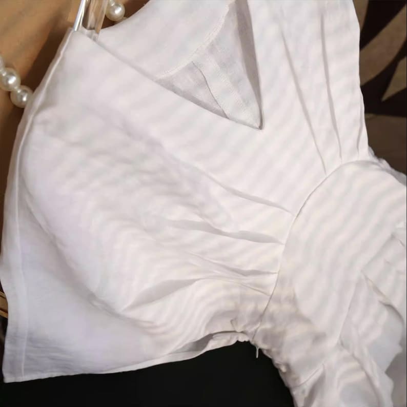 May include: A white, short-sleeved blouse with a V-neckline. The fabric has a subtle striped pattern. The blouse is hanging on a clear hanger, with a pearl necklace visible in the background.