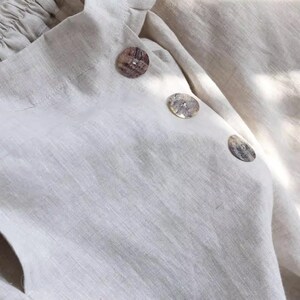 May include: Close-up of a cream-colored linen top with three decorative buttons. The buttons are round and have a mottled, natural appearance. The fabric has a textured weave and a soft, neutral tone.
