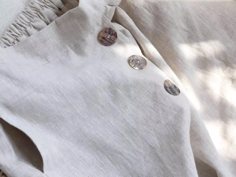 May include: Close-up of a cream-colored linen top with three decorative buttons. The buttons are round and have a mottled, natural appearance. The fabric has a textured weave and a soft, neutral tone.