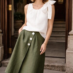May include: A sleeveless white top with a bow detail on the shoulder is paired with an olive green wrap skirt. The skirt features three large white buttons. The outfit is suitable for casual or semi-formal wear.