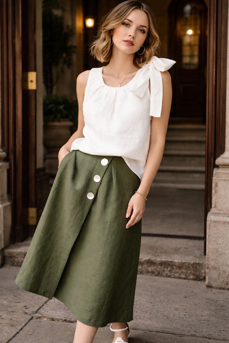 May include: A sleeveless white top with a bow detail on the shoulder is paired with an olive green wrap skirt. The skirt features three large white buttons. The outfit is suitable for casual or semi-formal wear.