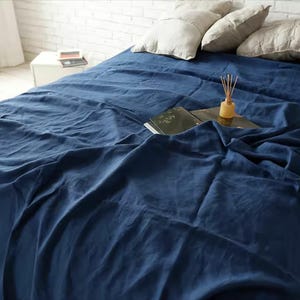 May include: A navy blue linen bed sheet covers a bed, with pillows and a book visible. The sheet is draped, creating soft folds and shadows. A small wooden diffuser sits on the book.