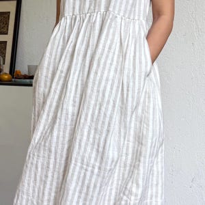 May include: A sleeveless midi dress featuring a vertical striped pattern in beige and white. The dress has a round neckline, a gathered waistline that creates a full skirt, and side pockets. The fabric has a textured appearance throughout the garment.