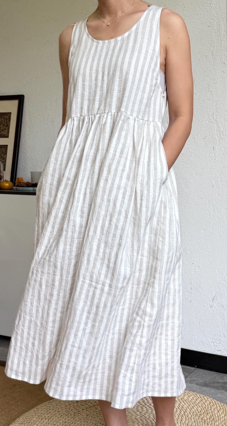 May include: A sleeveless midi dress featuring a vertical striped pattern in beige and white. The dress has a round neckline, a gathered waistline that creates a full skirt, and side pockets. The fabric has a textured appearance throughout the garment.