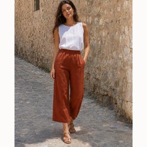 May include: A sleeveless white top with a round neckline paired with wide-leg rust-coloured trousers featuring an elasticated waistband. The trousers fall to a cropped length above the ankles, and the outfit is completed with brown strappy sandals.