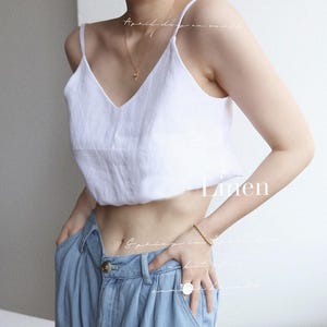 Könnte beinhalten: Weißes Leinen-Camisole-Top mit V-Ausschnitt und Spaghettiträgern. Das Wort "Linen" ist auf dem Bild aufgedruckt. Das Model trägt eine goldene Halskette, ein Armband und einen Ring. Das Model trägt hellblaue Jeans.