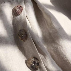 May include: Close-up of a beige linen fabric with three large, round buttons. The buttons are a mottled brown and tan color, and the fabric has a subtle texture. The buttons are sewn onto a placket.