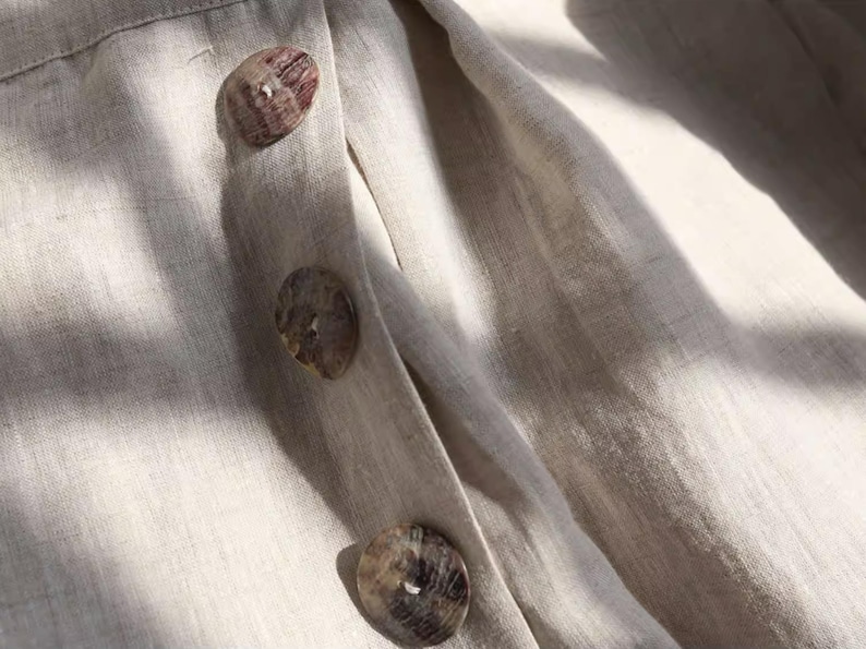 May include: Close-up of a beige linen fabric with three large, round buttons. The buttons are a mottled brown and tan color, and the fabric has a subtle texture. The buttons are sewn onto a placket.