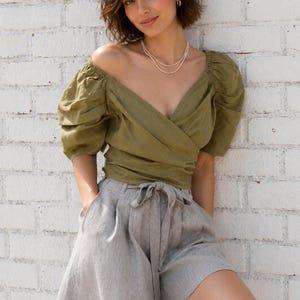 May include: An olive green wrap top featuring a deep V-neckline and voluminous puff sleeves paired with light grey high-waisted shorts that include a matching fabric tie belt at the waist and side pockets for a functional and structured look.