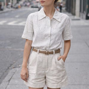 May include: A woman in a light linen shorts set, featuring a striped shirt and a woven belt. She wears sunglasses and has her hair up. The outfit is suitable for warm weather. The shorts and shirt are in neutral tones.
