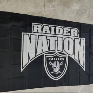 May include: Black and white flag with the text "Raider Nation" in a bold white font. The Raiders logo is in the lower center of the flag, featuring a silver helmet with a black face mask and crossed swords.