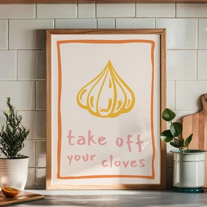 以下が含まれることがあります： 黄色のニンニクのイラストと「take off your cloves」というピンクの文字が書かれた、額縁に入ったキッチンプリントです。