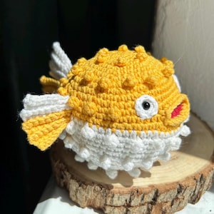 May include: A handmade crochet pufferfish toy. The fish features a yellow body with white and yellow fins and a white belly. The fish has a red mouth, a white eye with a black pupil, and a textured surface. The toy is sitting on a wooden slice.