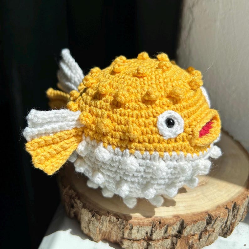 May include: A handmade crochet pufferfish toy. The fish features a yellow body with white and yellow fins and a white belly. The fish has a red mouth, a white eye with a black pupil, and a textured surface. The toy is sitting on a wooden slice.