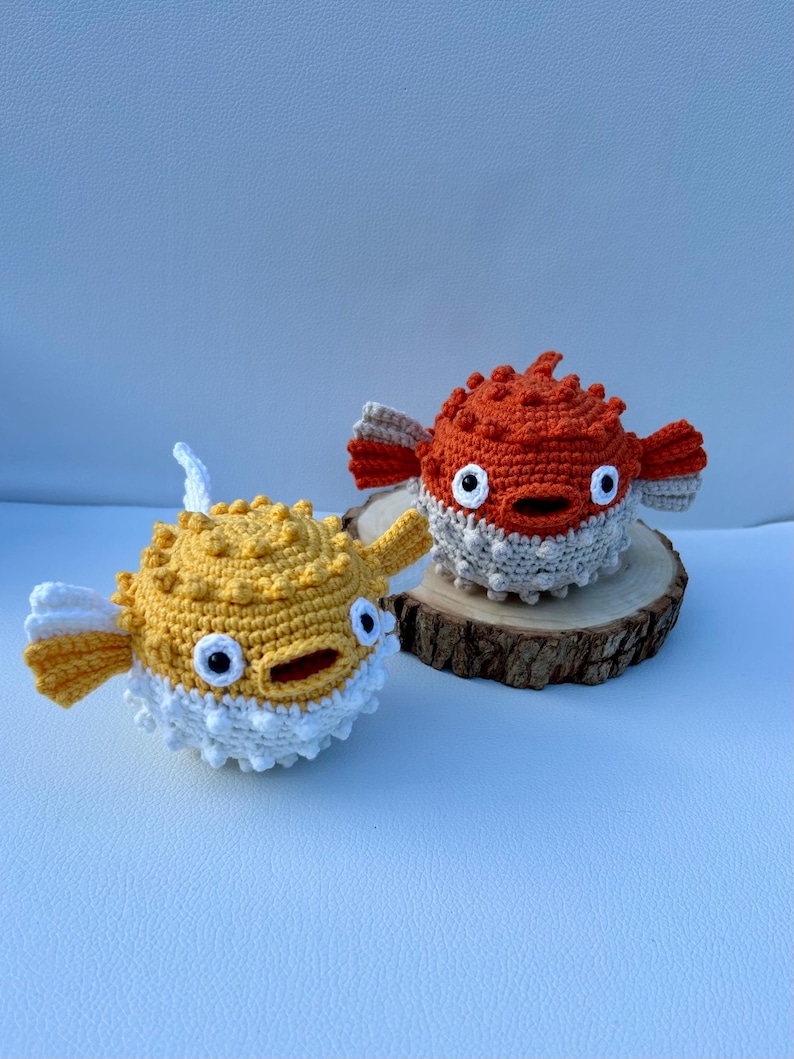 May include: Two handmade crochet pufferfish toys. One is yellow and white, the other is orange and white. Both have large, round eyes and are sitting on a wooden slice. The toys are made with a textured stitch.