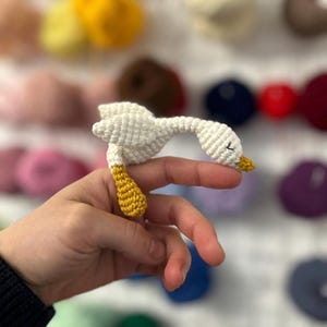 May include: A small white crocheted goose with yellow feet and a beak. The goose is being held by a hand.