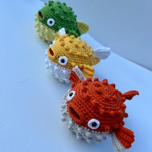 May include: Three handmade crochet pufferfish toys in a row. The top one is green, the middle one is yellow, and the bottom one is orange. Each has white and beige accents, black and white eyes, and a red mouth. The toys are soft and textured.