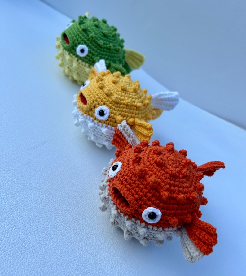 May include: Three handmade crochet pufferfish toys in a row. The top one is green, the middle one is yellow, and the bottom one is orange. Each has white and beige accents, black and white eyes, and a red mouth. The toys are soft and textured.