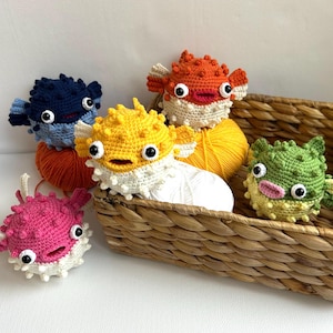 May include: Five crocheted pufferfish toys in different colours, including blue, yellow, orange, pink, and green. The toys are sitting in a wicker basket with balls of yarn.