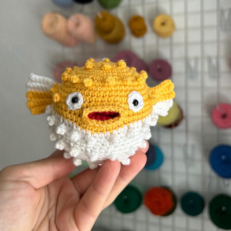May include: A handmade crochet pufferfish toy. The toy is primarily yellow with white and red accents. It has black eyes and white fins. The pufferfish has a textured, spiky appearance, and is held in a person's hand.