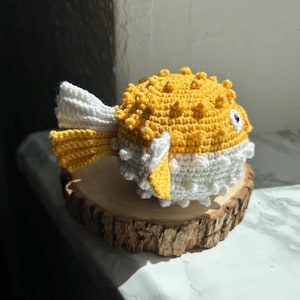 May include: A handmade crochet pufferfish toy in yellow and white, with a yellow tail and fins. The toy is sitting on a round, wooden slice. The pufferfish has a textured, spiky appearance.