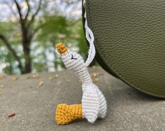 Può includere: Un portachiavi a forma di oca bianca all'uncinetto con piedi gialli, appeso a una borsa di pelle verde.