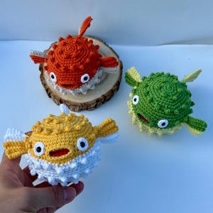 May include: Three handmade crochet pufferfish in orange, yellow, and green. The orange fish rests on a wooden slice. The yellow fish is held in a hand. Each fish has white accents, black eyes, and a smiling mouth.