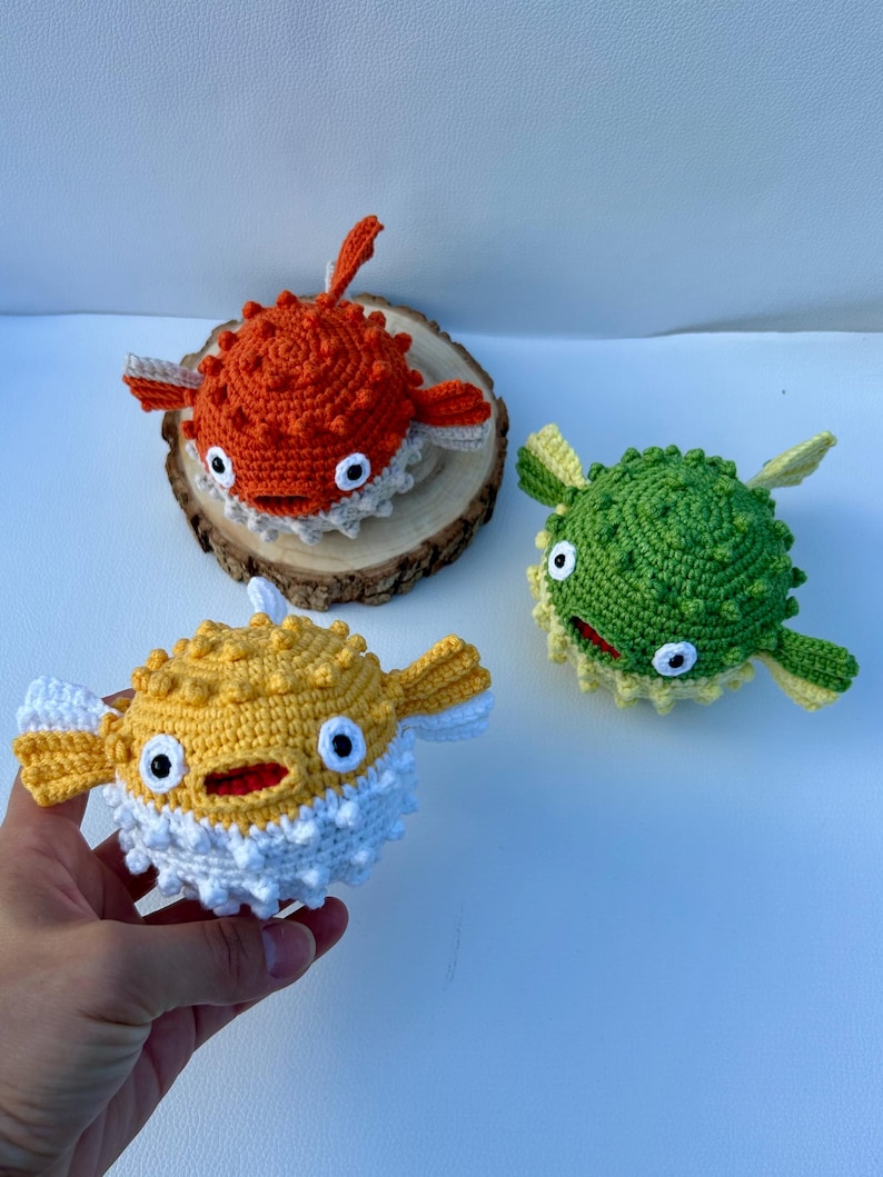 May include: Three handmade crochet pufferfish in orange, yellow, and green. The orange fish rests on a wooden slice. The yellow fish is held in a hand. Each fish has white accents, black eyes, and a smiling mouth.